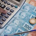 Pessoa usando calculadora ao lado de notas de real e moedas brasileiras, representando cálculo financeiro, controle de despesas e planejamento de dinheiro.